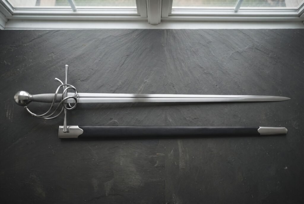 Hand forged swept hilt rapier with a coiled polished steel guard and wire wrapped grip laid on a dark slate slab beside its black leather scabbard