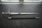 Hand forged swept hilt rapier with a coiled polished steel guard and wire wrapped grip laid on a dark slate slab beside its black leather scabbard