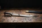 Hand forged talwar sword with a mirror polished curved blade, blackwood grip and disc pommel on a scarred dark timber workbench beside its black leather scabbard