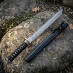 Hand forged tanto with cord wrapped handle and black lacquered saya lying diagonally across a lichen covered boulder with leaves