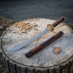 Hand forged tanto sword with a brown cord grip and round brass tsuba resting on a sawn tree stump beside its brown leather wrapped saya and wood shavings