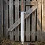 Hand forged two handed kriegsmesser with a broad curved single edged blade and long grip leaning against a weathered timber gate beside its leather scabbard