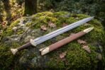 Hand forged Ulfberht sword with polished high carbon steel blade, brass lobed pommel and tan leather scabbard, laid on a moss covered granite boulder outdoors
