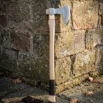 Hand forged viking battle axe with a bearded carbon steel head and pale ash haft, standing upright against a weathered red brick wall in daylight
