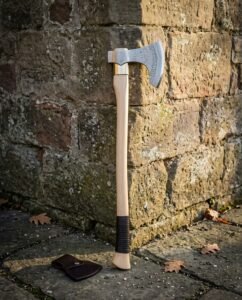 Hand forged viking battle axe with a bearded carbon steel head and pale ash haft, standing upright against a weathered red brick wall in daylight