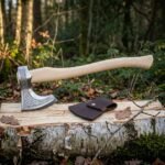 Hand forged viking bearded axe with an etched Norse interlace steel head and light birch haft, resting on a split birch log beside its dark leather sheath