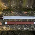 Hand forged Viking broadsword with wire wrapped grip, brass fittings and brown leather scabbard laid flat on mossy flagstone and photographed from above