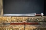 Hand forged viking rune sword with a broad blade carrying a darkened Elder Futhark inscription, red leather grip and brown scabbard on a sandstone ledge