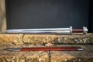 Hand forged viking rune sword with a broad blade carrying a darkened Elder Futhark inscription, red leather grip and brown scabbard on a sandstone ledge