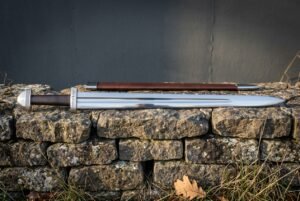 Hand forged Viking sword in polished high carbon steel with brown leather scabbard and brazil nut pommel, laid across a low drystone wall in daylight