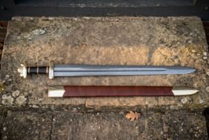 Hand forged Viking sword with a bright polished high carbon steel blade and brown leather scabbard laid across a warm sandstone ledge in daylight