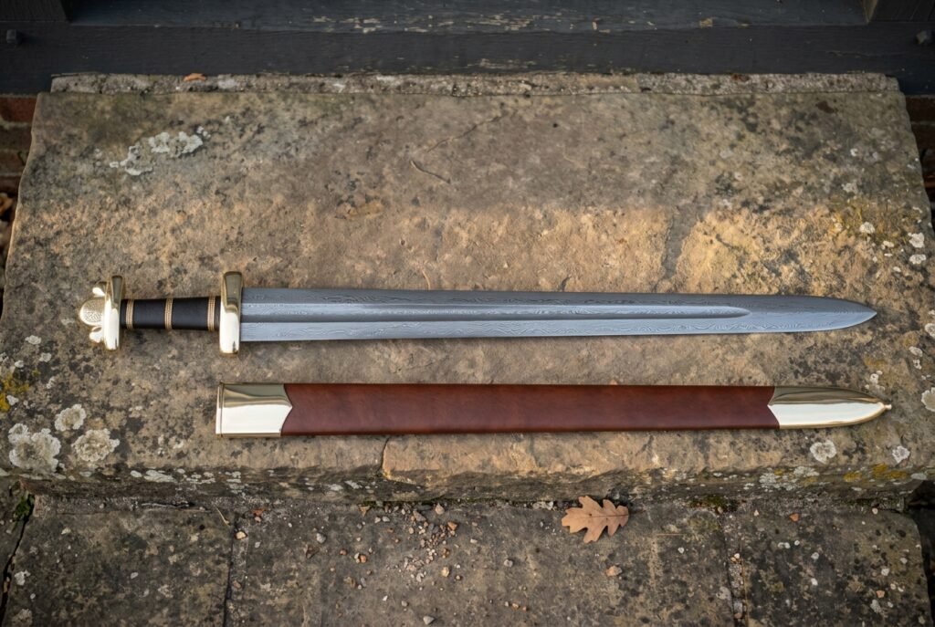 Hand forged Viking sword with scabbard in brown leather and a lobed steel pommel, laid on a sandstone step beside its belt in daylight