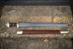 Hand forged Viking sword with scabbard in brown leather and a lobed steel pommel, laid on a sandstone step beside its belt in daylight