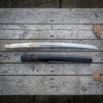 Hand forged wakizashi sword with a polished bone handle and brass tsuba laid on weathered grey timber decking beside its dark lacquered wooden saya