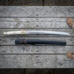 Hand forged wakizashi sword with a polished bone handle and brass tsuba laid on weathered grey timber decking beside its dark lacquered wooden saya