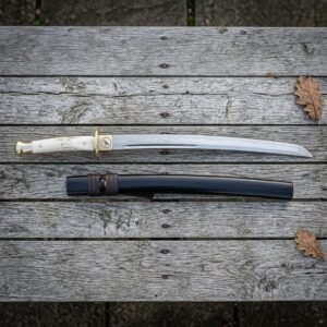 Hand forged wakizashi sword with a polished bone handle and brass tsuba laid on weathered grey timber decking beside its dark lacquered wooden saya