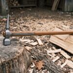 Hand forged war hammer with a polished steel square face and long rear spike on a hardwood haft resting on a tree stump among split firewood