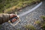 Hand gripping the black horn grip and rune engraved brass knuckle bow of a curved Damascus steel szabla over a gravel path