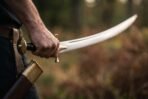Hand holding a curved talwar sword by its black grip and brass cross guard outdoors, brass topped scabbard at the hip