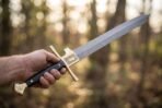 Hand holding a Damascus falchion by its black grip with engraved brass guard plate and brass pommel in an autumn forest