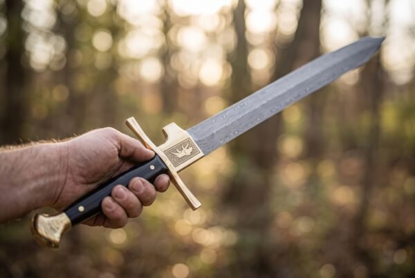 Hand holding a Damascus falchion by its black grip with engraved brass guard plate and brass pommel in an autumn forest