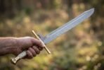 Hand holding a Damascus langes messer by its stag horn grip with a brass cross guard against a soft autumn forest