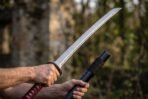 Hands drawing a short Japanese sword with red cord wrapped handle from its black lacquered saya outdoors