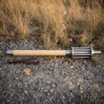 High carbon steel bar mace with pale ash haft and steel ferrules lying on gravel beside a brown leather wrist strap
