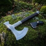 High carbon steel bearded axe with a rune etched head, black wrapped haft and steel butt cap, laid on a mossy rock beside a leather sheath