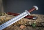 High carbon steel broadsword with a rune engraved blade, russet leather grip and lobed pommel, laid on a stone wall beside its scabbard