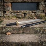 High carbon steel dao sword with a black cord wrapped grip, brass guard and brass pommel, laid on a stone ledge beside its black scabbard