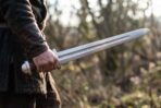 High carbon steel falchion with wooden grip and steel pommel held low in one hand against a wintry woodland background