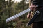High carbon steel grosse messer held by the grip in a woodland clearing with its black leather sheath hanging at the hip