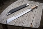 High carbon steel grosse messer with knotwork etched blade and pale wood grip laid beside its black leather sheath on a table