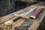 High carbon steel longsword with a brass disc pommel, black leather grip and curved brass guard, laid on a stone wall by its red scabbard