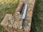 High carbon steel messer sword with a dark wooden grip, steel cross guard and side nagel, laid on a brick wall beside its leather sheath