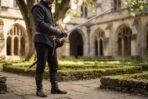 High carbon steel musketeer sword with a long slim blade and swept cup hilt held by a man in a black doublet in a cloister courtyard