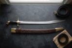 High carbon steel scimitar with a black grip, brass cross guard and hooked brass pommel, on a dark table with its tooled leather scabbard