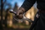 High carbon steel short sword with a faceted steel pommel and looped side ring guard held in a gloved hand, black scabbard at the hip