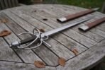 High carbon steel side sword with a swept steel hilt, dark wrapped grip and round pommel, laid on a wooden garden table by its brown scabbard