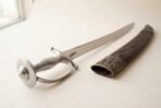 High carbon steel talwar with all steel hilt and domed disc pommel lying beside its tooled metal mounted scabbard