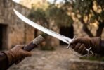 High carbon steel talwar with all steel hilt held out by gloved hands in a sunlit stone courtyard with olive trees