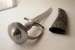 High carbon steel talwar with disc pommel and knuckle guard, close up of the hilt beside the tooled scabbard