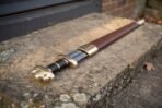 High carbon steel Viking sword with a brass lobed pommel and guard and black grip, resting on a stone step in its brown leather scabbard