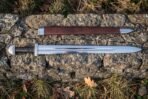 High carbon steel Viking sword with brown leather grip and steel guard laid on a stone wall below its brown leather scabbard