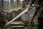 High carbon steel Viking sword with fullered blade and rounded steel pommel held point down beside a brown leather scabbard