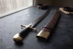 Close up of the hooked brass pommel and black grip of a scimitar beside its tooled leather scabbard with a brass throat on a dark table