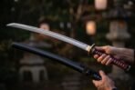 Katana with crimson cord wrapped handle and brass tsuba being drawn from a black lacquered saya in an evening garden