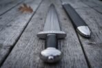 Close up of a lobed steel pommel and rune engraved guard on a Damascus sword, black scabbard with a steel chape laid alongside on grey boards