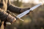 Longsword with a bright fullered blade held flat across two gloved hands, brass guard and disc pommel to one side, field behind